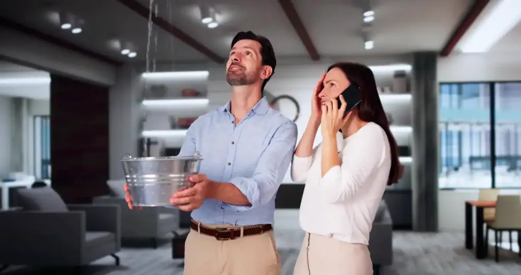 A man catches a ceiling leak in a bucket as a woman calls Water Damage Restoration Milwaukee County, WI.